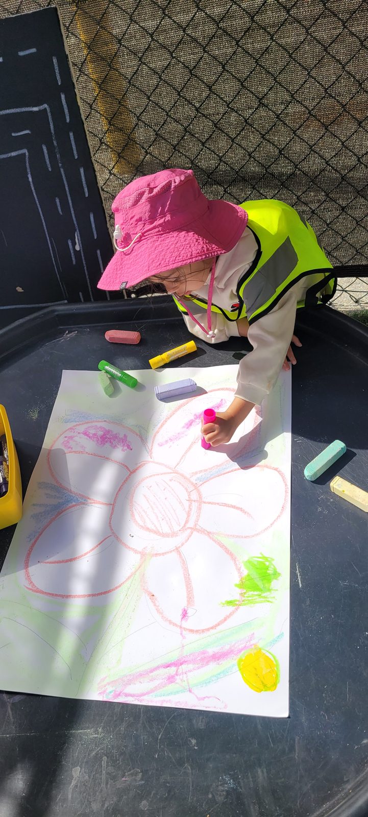 A child drawing a large flower with oil pastels at an outdoor table